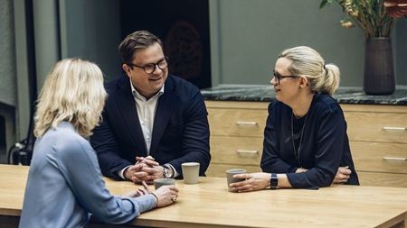 Tre personer samtalar vid ett träbord med koppar och en grön vägg i bakgrunden.
