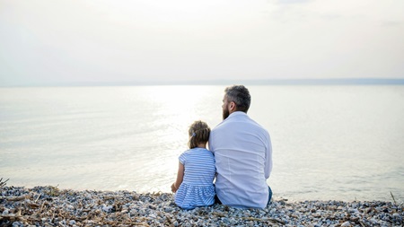 En man och en liten flicka sitter bredvid varandra på en stenig strand och tittar ut över ett lugnt, solbelyst hav under en ljus himmel.