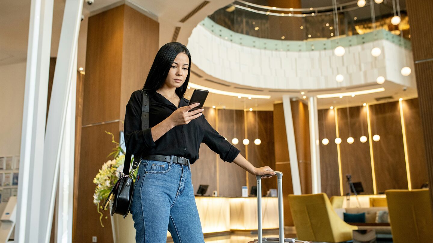 Kvinna med resväska står i en hotellobby och tittar på sin mobil.