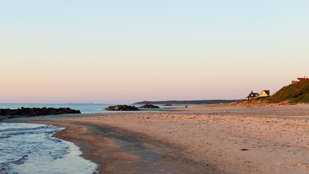 Strand med sand, vatten, klippor och hus i fjärran under solnedgång eller soluppgång