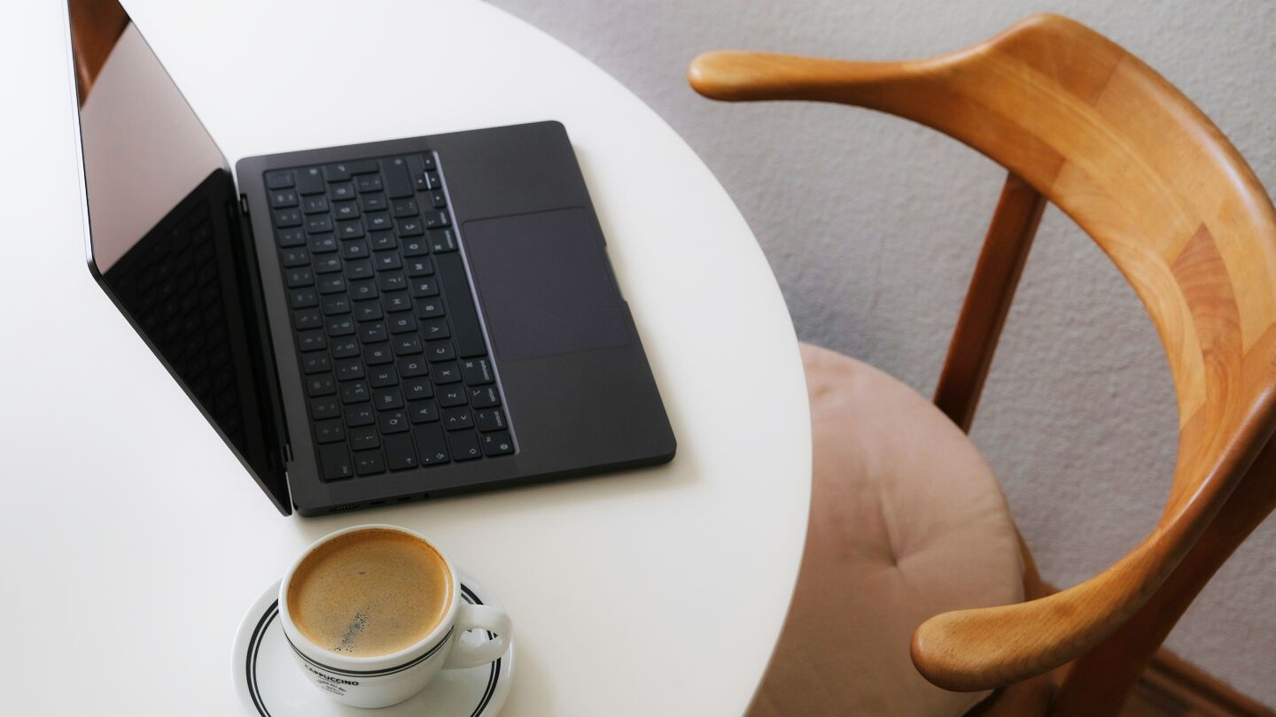 En öppen laptop på ett vitt runt bord bredvid en kopp kaffe, med en stol i trä och stoppad sits intill.