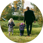 En vuxen som går utomhus på gräset med två barn, omgiven av grönska och träd.