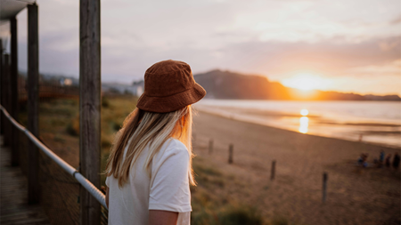 Kvinna med hatt ser ut över stranden vid solnedgång.