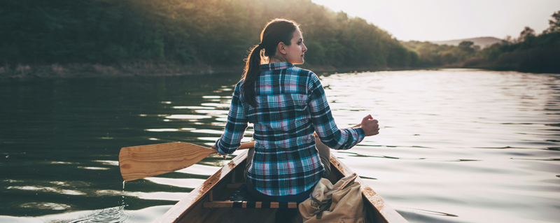 En person sitter i en träkanot och paddlar på en lugn sjö omgiven av grönskande kullar. Solen lyser svagt i bakgrunden och en ryggsäck ligger i kanoten.